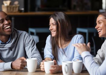 Attain on 5th apartment homes with Four friends sit at a table, smiling and laughing together while holding white mugs.