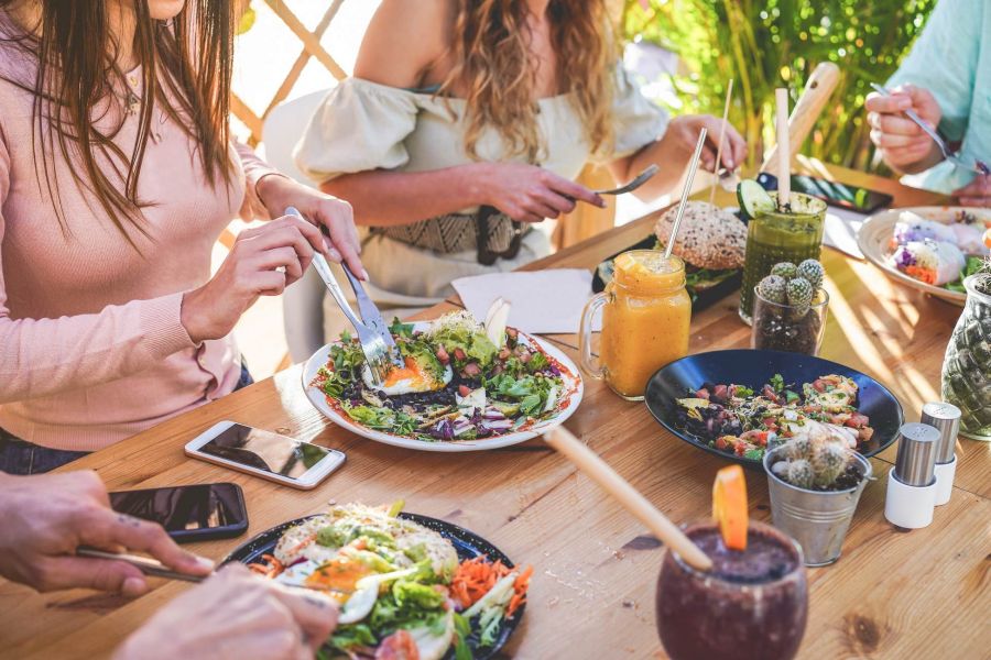 Attain at Chic's Beach Three women eating salads and drinks at an outdoor wooden table with phones and cutlery visible.