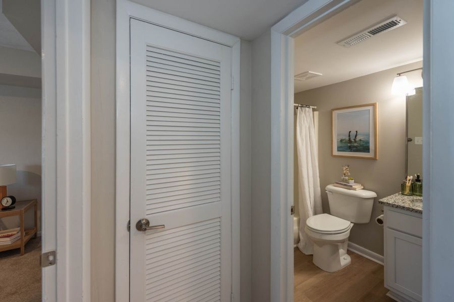 Attain at Chic's Beach homes with Hallway view of a bathroom with a toilet, vanity, and shower curtain, next to a closed louvered closet door.
