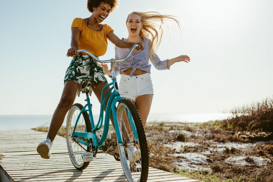Attain on 5th apartment homes with Two young women laughing, riding a blue bicycle together on a boardwalk by the beach on a sunny day.