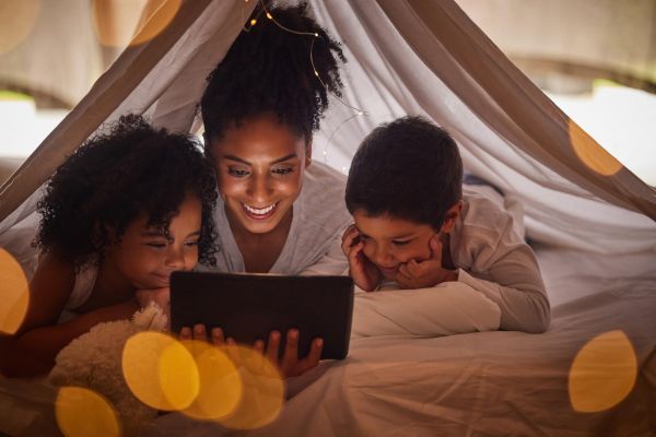 Attain on 5th apartment homes with A woman and two children smile while watching a tablet together inside a cozy blanket fort with soft lights.