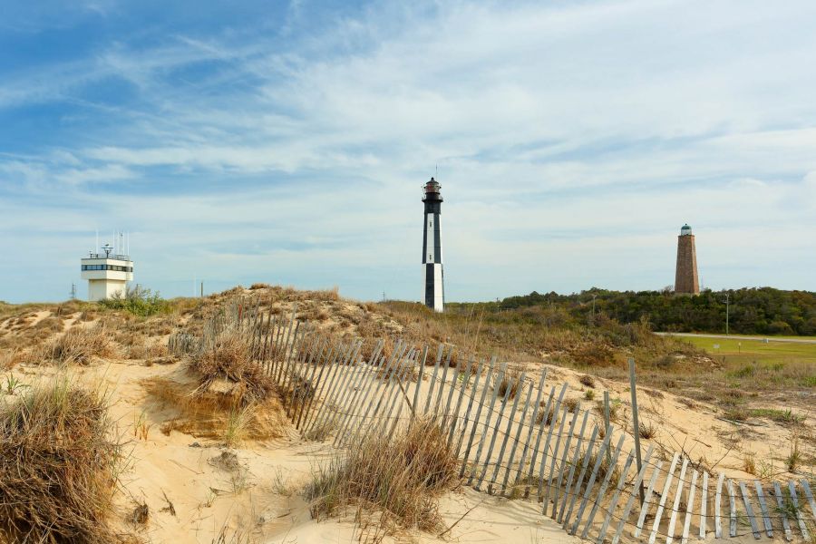 Attain at Chic's Beach with A sandy dune with grass, a wooden fence, and two lighthouses in the distance under a partly cloudy sky.