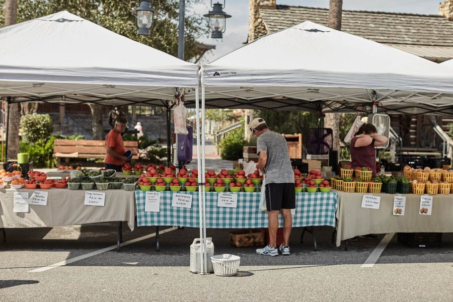 Attain at Chic's Beach with People browse fresh produce under white tents at an outdoor farmers market on a sunny day.