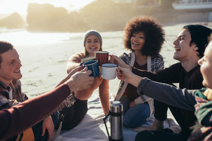 Attain on 5th apartment homes with Five friends smiling and toasting mugs together while sitting on a blanket at the beach.